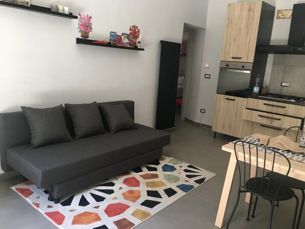 Een moderne woonkamer met een grijze bank, een kleurrijk vloerkleed en een keuken met houten kastjes en een eettafel.