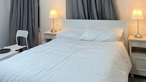 A neatly made bed with white bedding, two bedside lamps, a white chair, and grey curtains. - Impeccable Studio Apartment in West London (London)