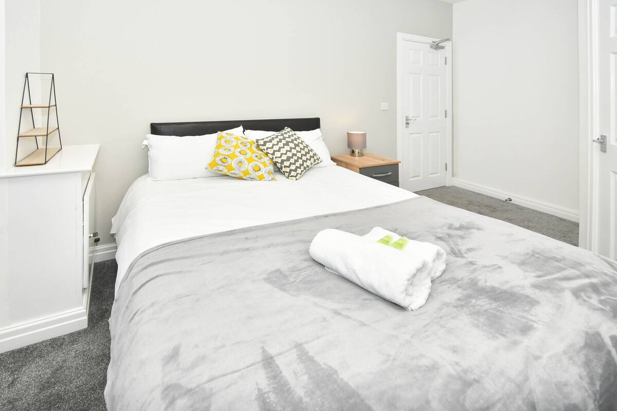 Una cama bien hecha con un edredón gris y dos almohadas, una mesita de noche con una lámpara y una puerta blanca en una habitación de estilo minimalista.