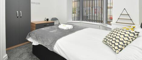 A bedroom with a bed, a wardrobe, a bedside table, a lamp, and a window with blinds.