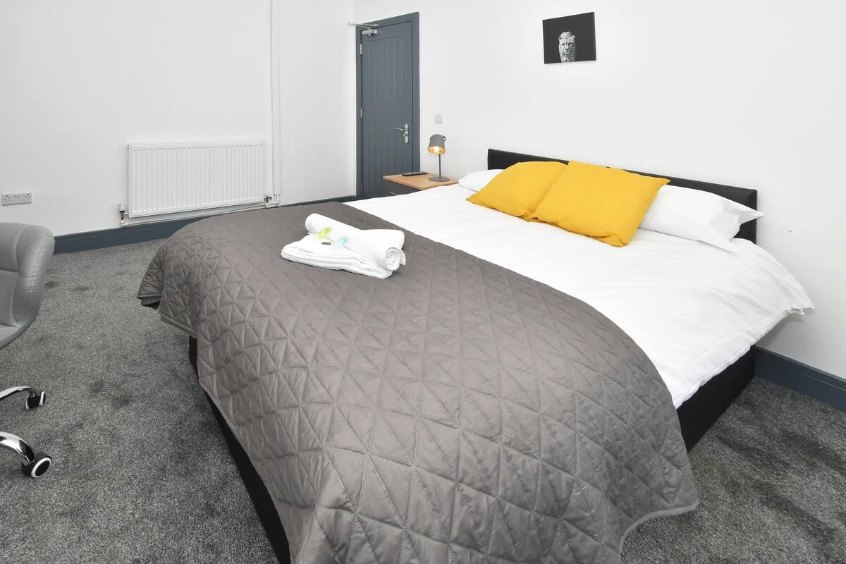 Un lit bien fait, recouvert d’une courtepointe grise matelassée, d’une couette blanche et d’un oreiller jaune. On aperçoit une serviette pliée sur le lit, une petite table de chevet avec une lampe, et une chaise dans le coin de la pièce.
