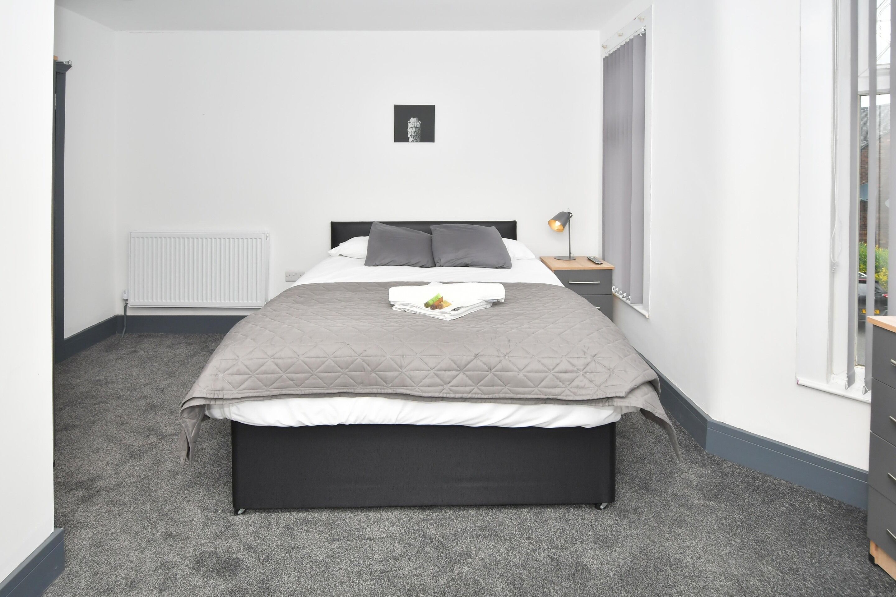 A neatly arranged bedroom with a bed, bedside table, lamp, and a framed picture on the wall.