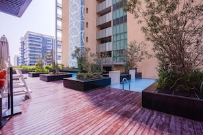 A rooftop pool area with wooden decking, planters with greenery, and a view of modern buildings in the background.