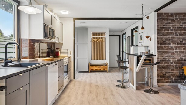 Une cuisine moderne avec un îlot central, un four à micro-ondes encastré et un évier. On aperçoit un banc en bois et un mur en briques à l’arrière-plan.