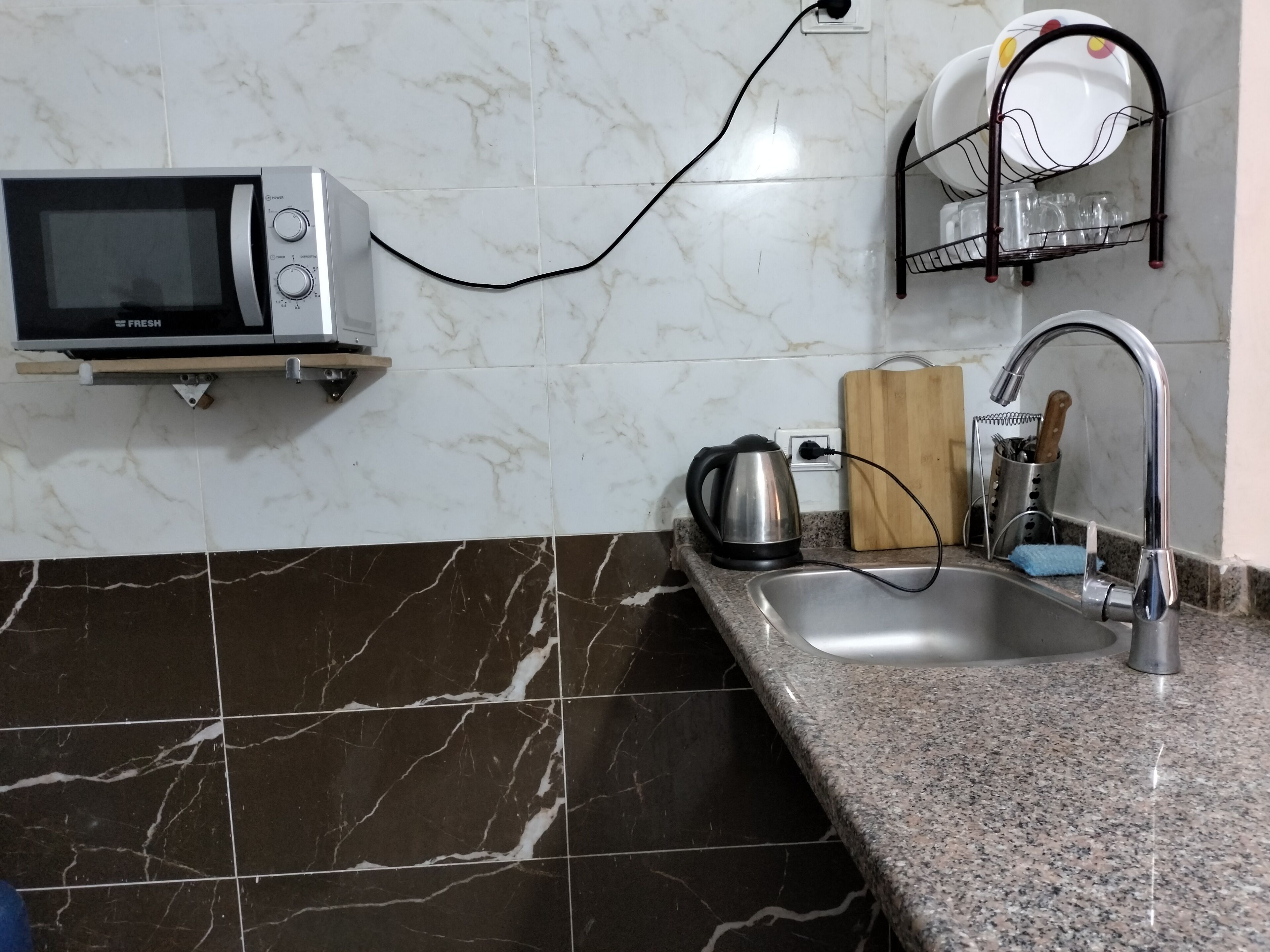 A kitchen with a microwave, kettle, sink, and dish rack.