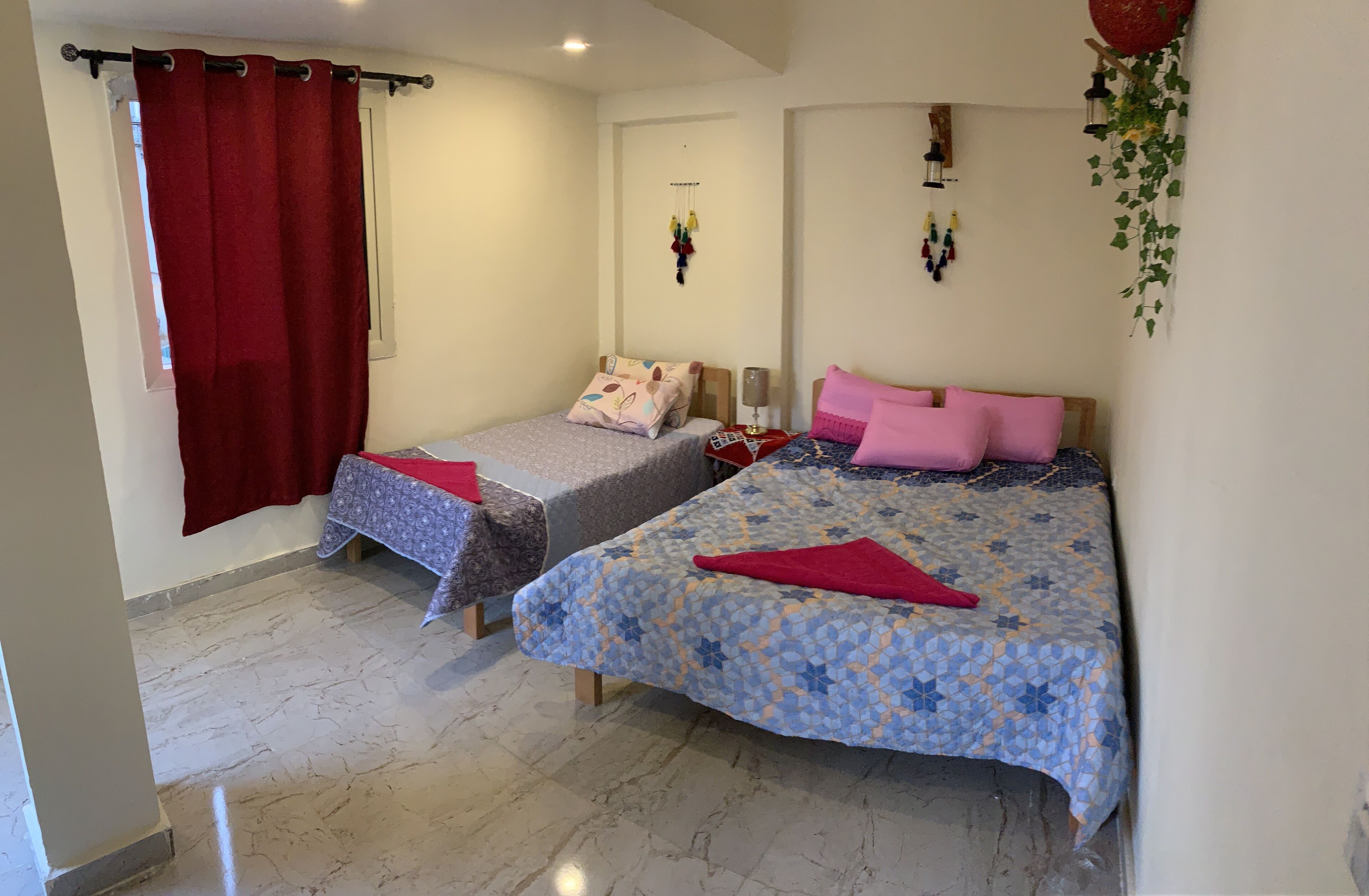 A bedroom with two beds, a window with red curtains, a wall-mounted light fixture, and a hanging plant.