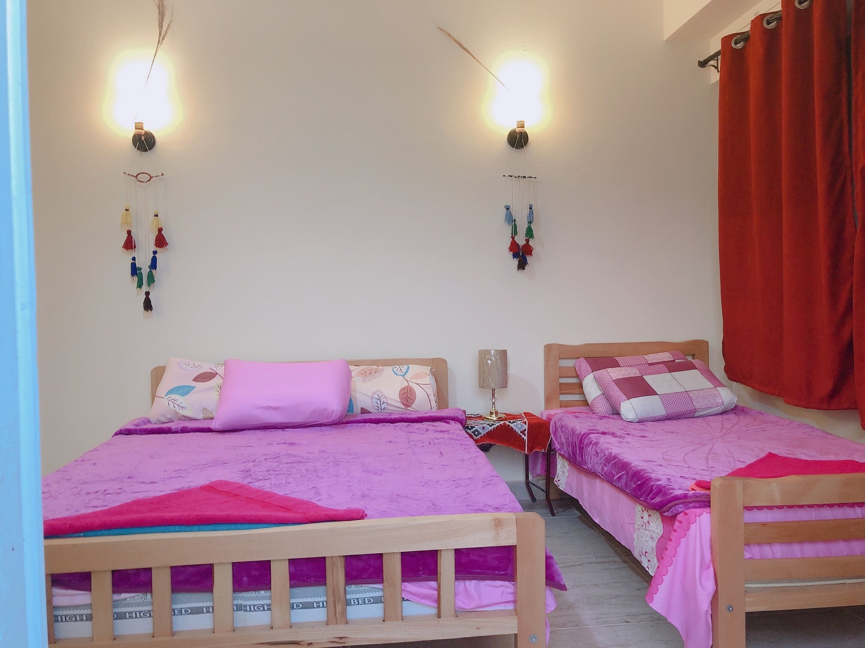 Una habitación con dos camas de madera, cada una con una colcha y almohadas rosas. Hay luces fijadas a la pared y un tapiz con flecos.
