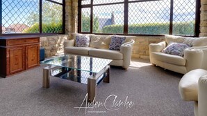 A spacious living room with a glass coffee table, two beige sofas with floral cushions, a wooden cabinet, and large windows offering a view of greenery and a distant building. - Stunning Residence set on Cliffside in Scarborough (Scarborough)