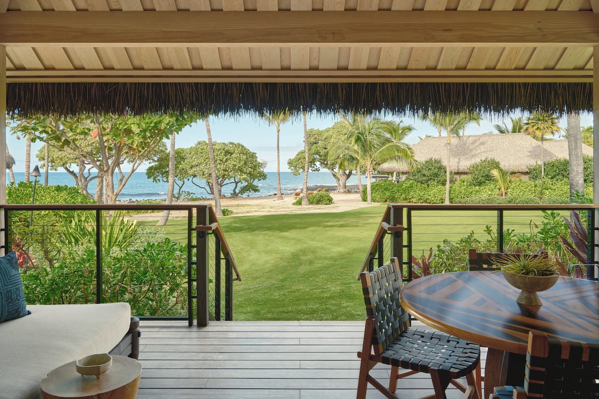 A wooden deck with a table and chairs, overlooking a well-maintained lawn and palm trees, with a view of the sea in the background.
