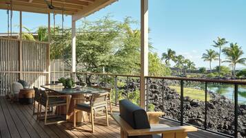 A wooden deck with a table and chairs, overlooking a rocky shoreline with palm trees.