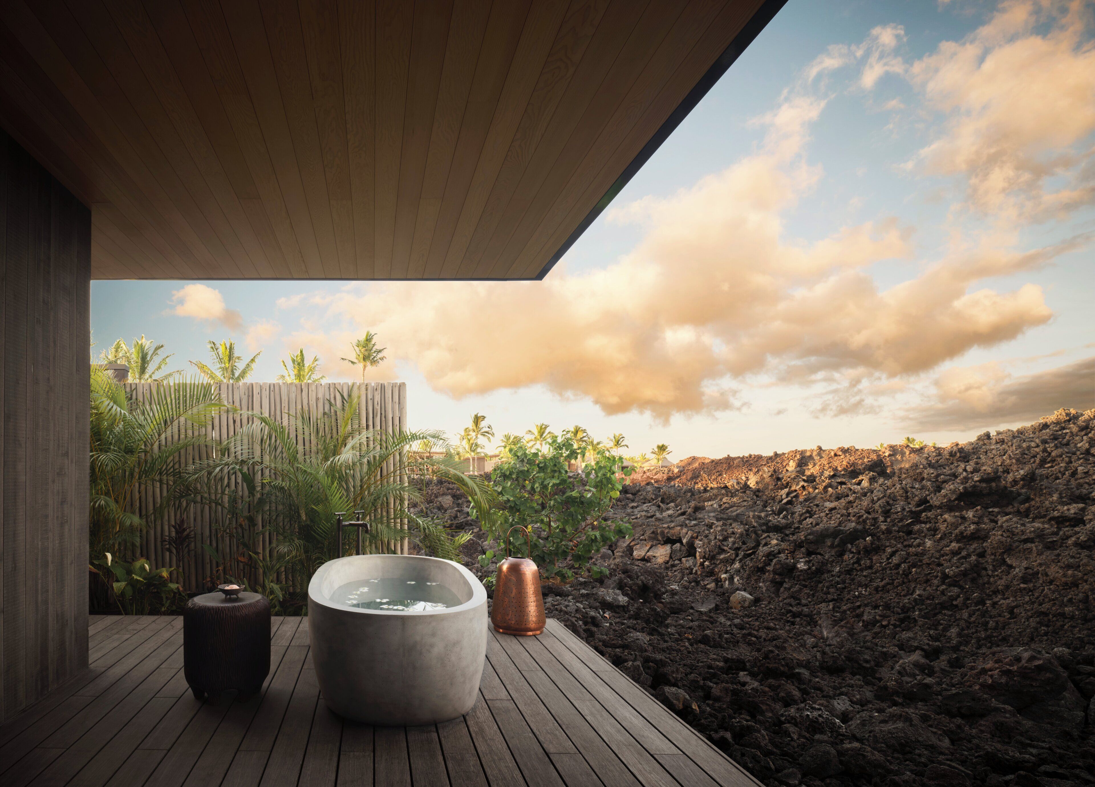 Una terraza de madera con un jacuzzi de piedra, un cerramiento de bambú y vistas a un paisaje volcánico.