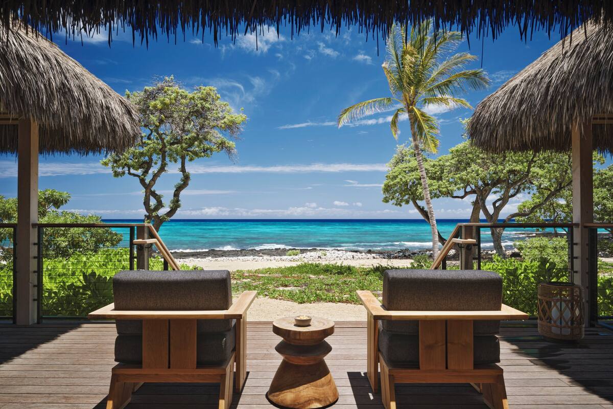 A wooden deck with two lounge chairs facing a beach with clear turquoise water and palm trees.