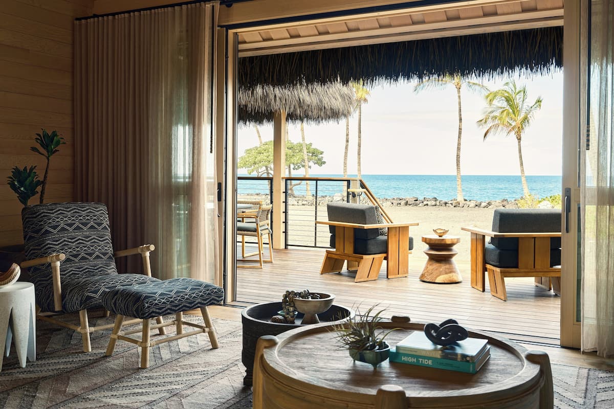 A room with a view of the beach, featuring a wooden deck, a chair, a table with books, and a plant.