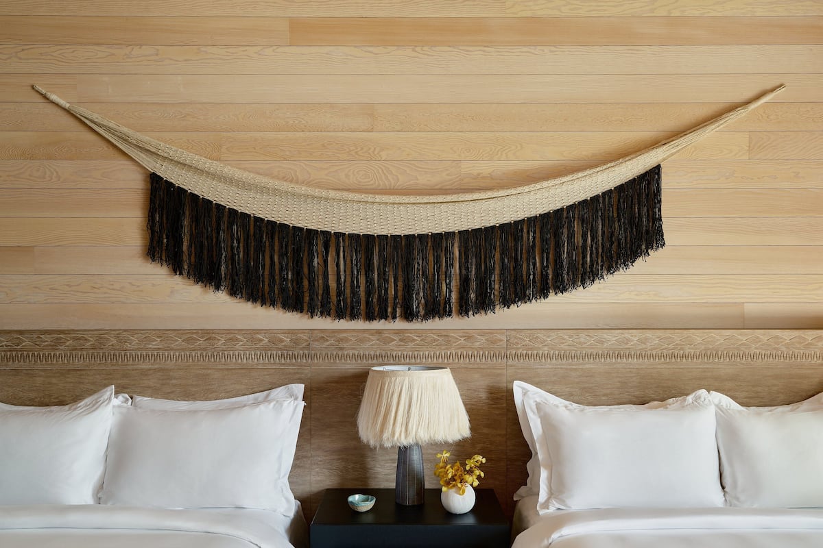 A neatly arranged bedroom with a wooden headboard, a woven wall hanging, a bedside lamp, and a small vase with flowers.