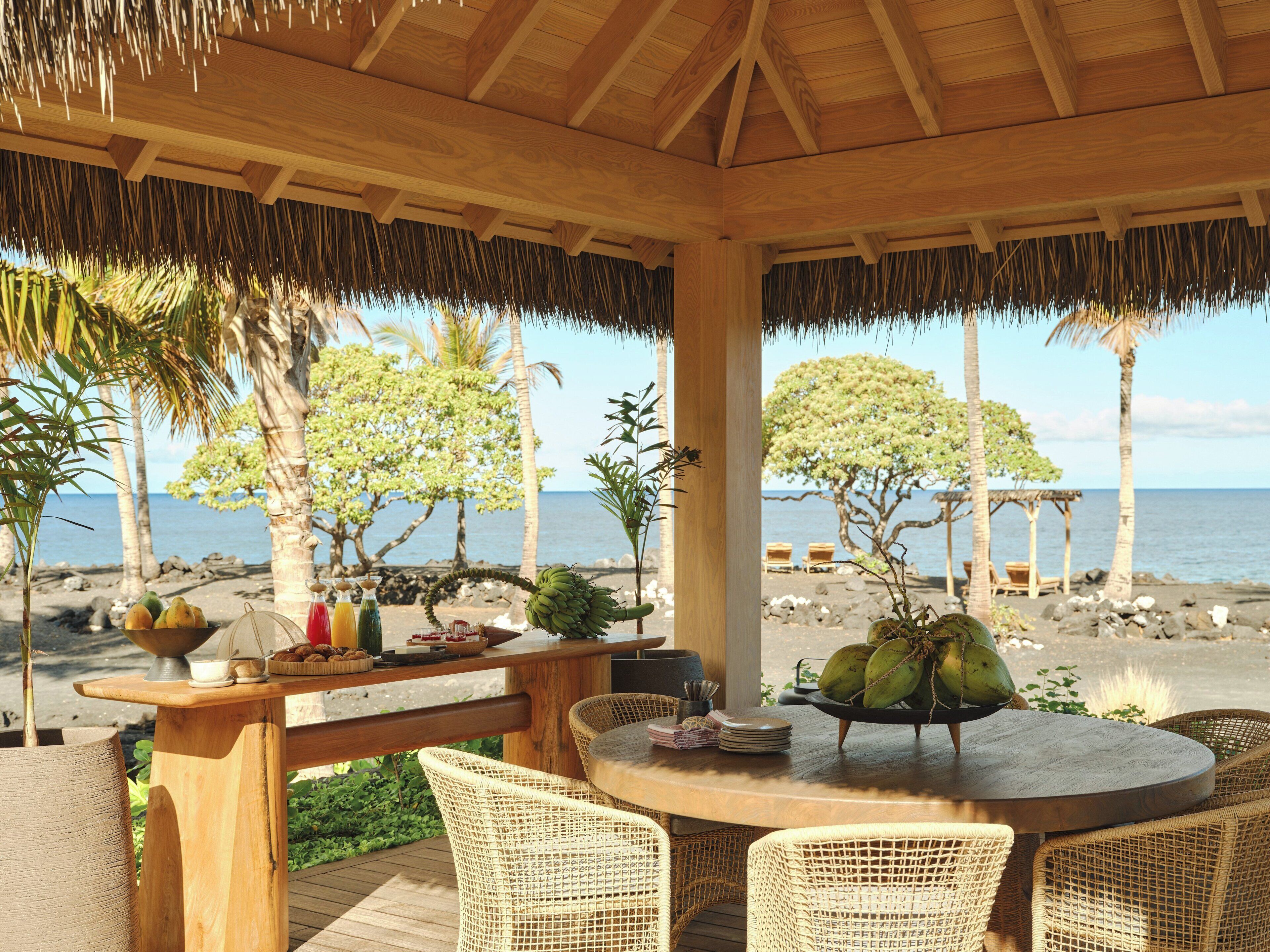 A wooden table with wicker chairs, a thatched roof, and a view of the sea and trees.