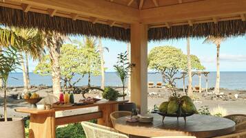 A wooden table with wicker chairs, a thatched roof, and a view of the sea and trees.