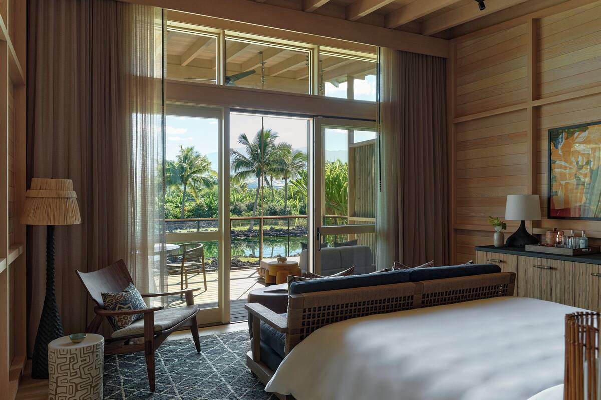 A hotel room with a large bed, a seating area with a chair and table, a view of a pool and palm trees, and a ceiling fan.