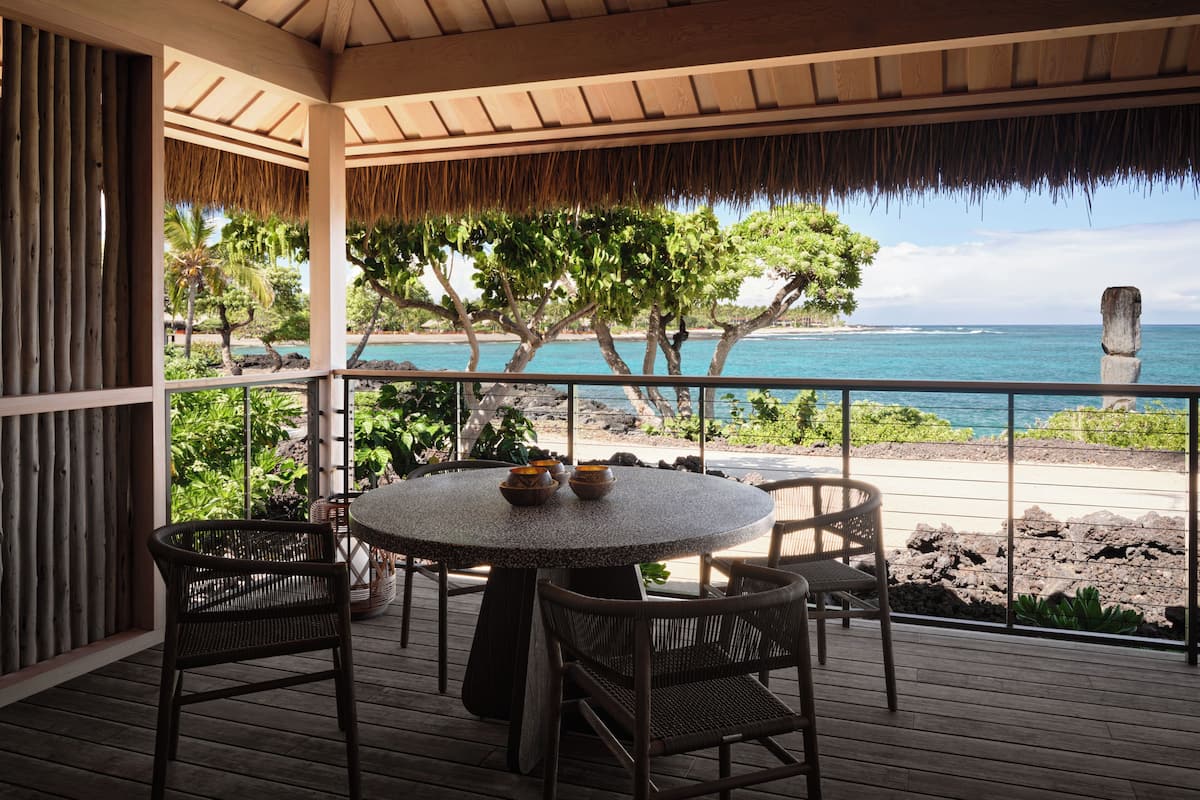 A wooden deck with a round table and wicker chairs, overlooking a beach with a clear view of the ocean and a stone headstone.