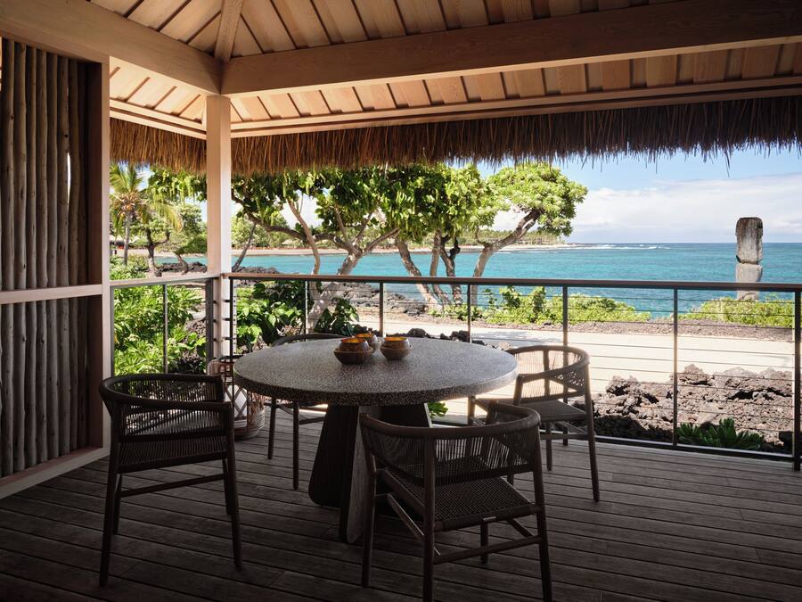 A wooden deck with a round table and wicker chairs, overlooking a beach with a clear view of the ocean and a stone headstone.
