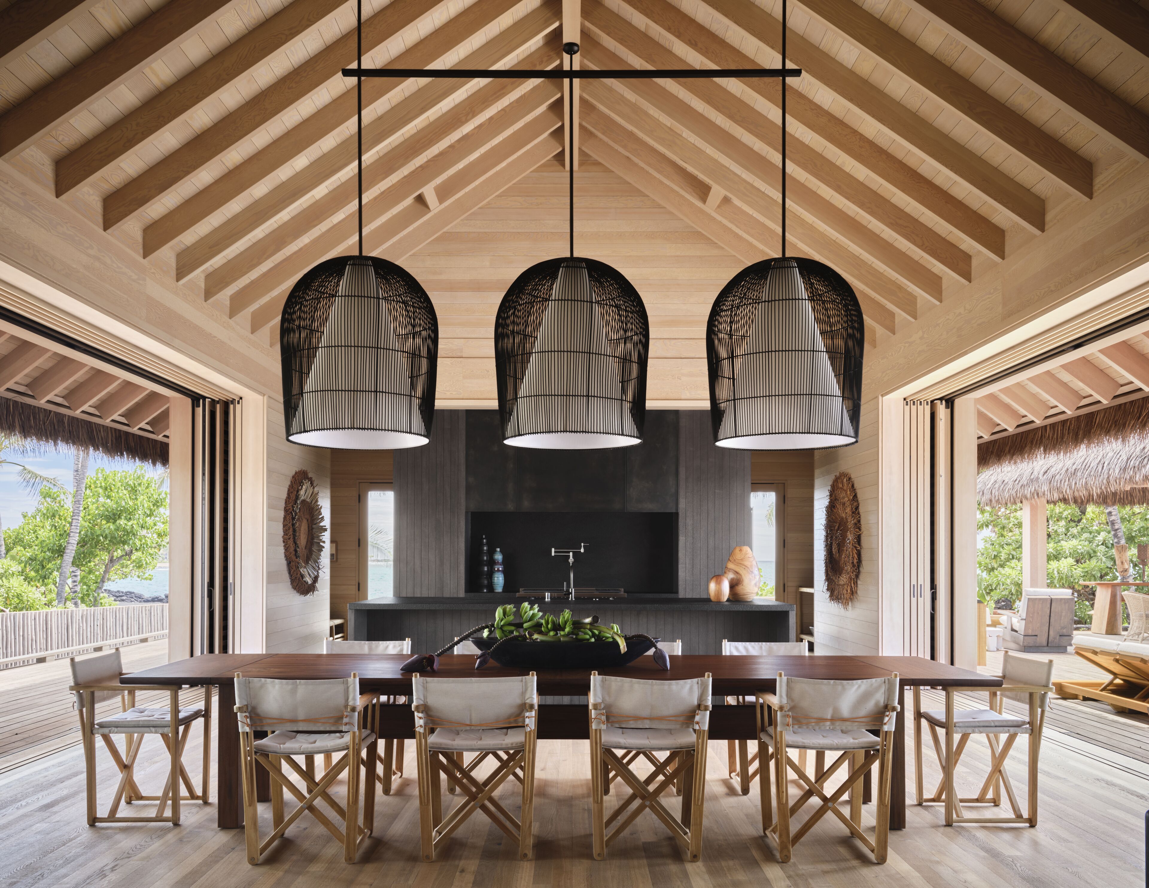 A spacious dining area with a long wooden table and chairs, a kitchen in the background, and a view of the outdoors.