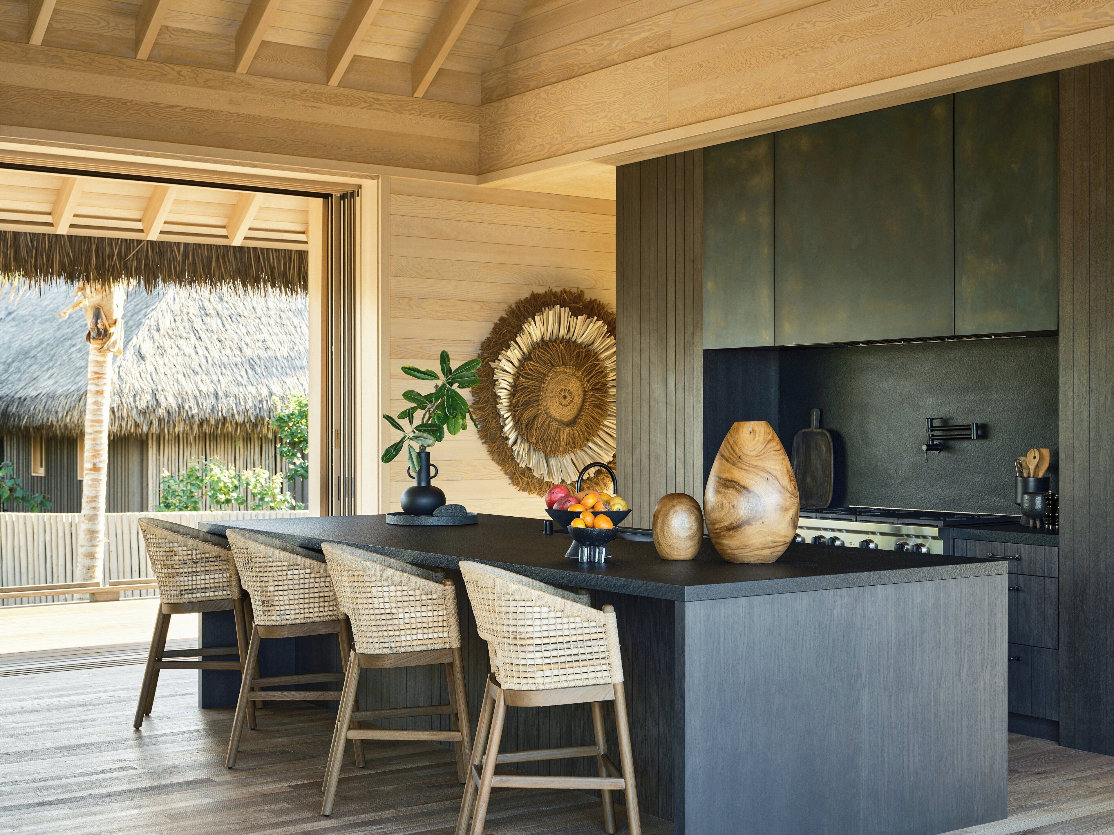 A modern kitchen with a large island, bar stools, a stove, and a dining area with a thatched roof view.