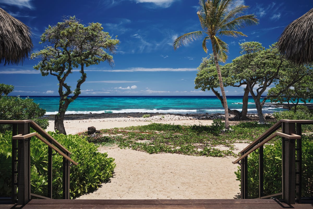 A beach view with a wooden staircase leading to a sandy path, surrounded by trees and thatched-roof structures.