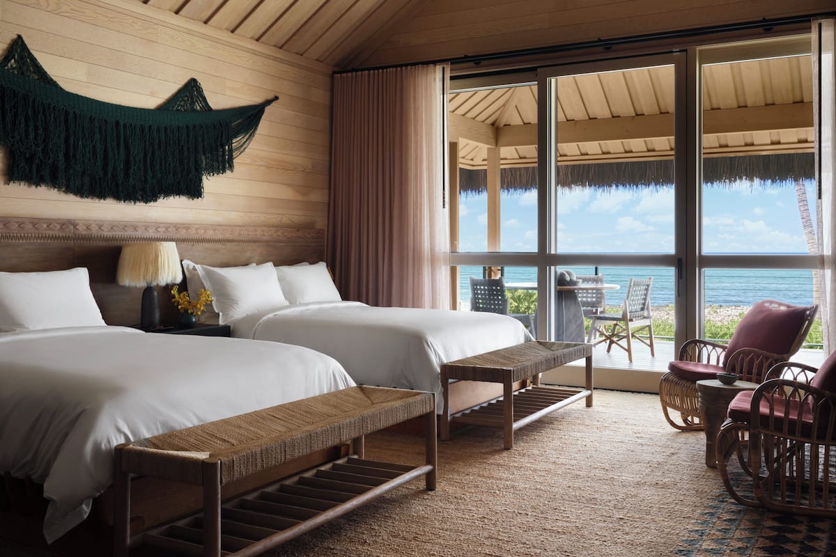 A room with two beds, a bench, wicker chairs, and a view of the ocean.