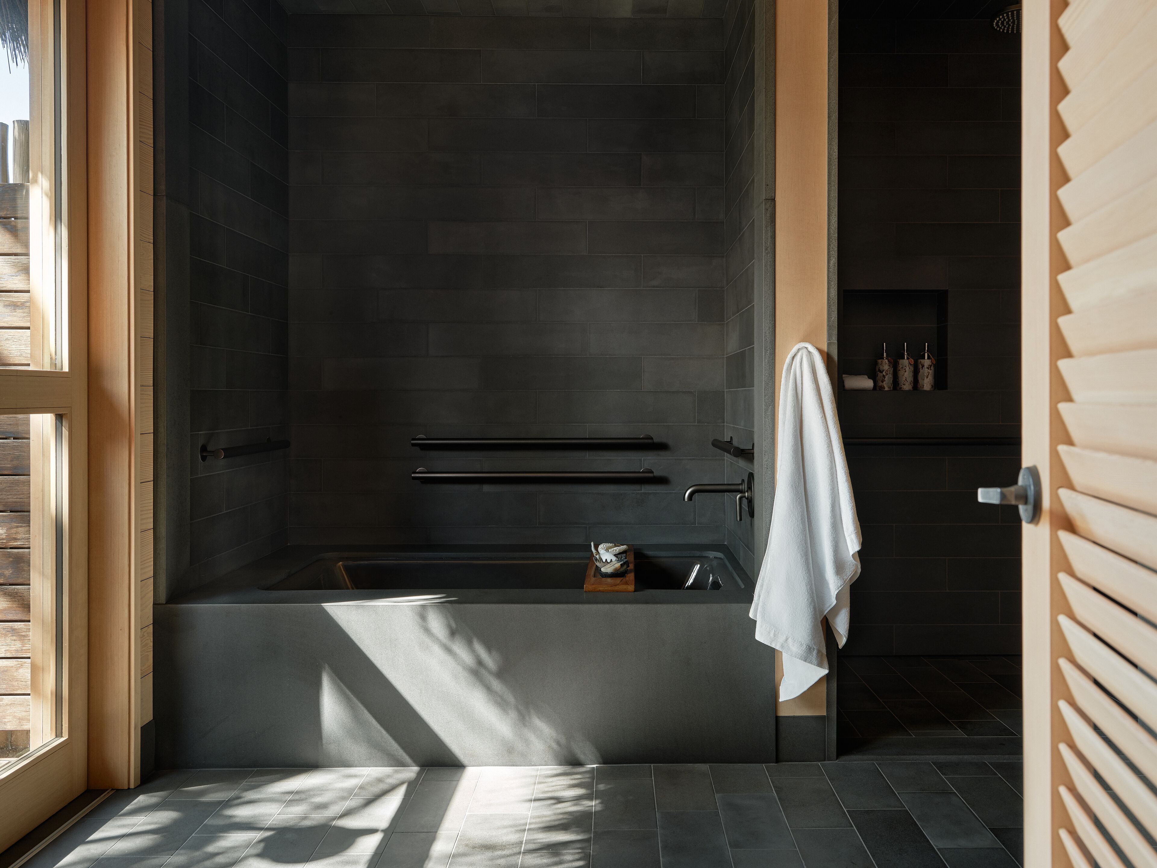Un baño moderno con una pared de azulejos oscuros, una bañera y una puerta de madera.