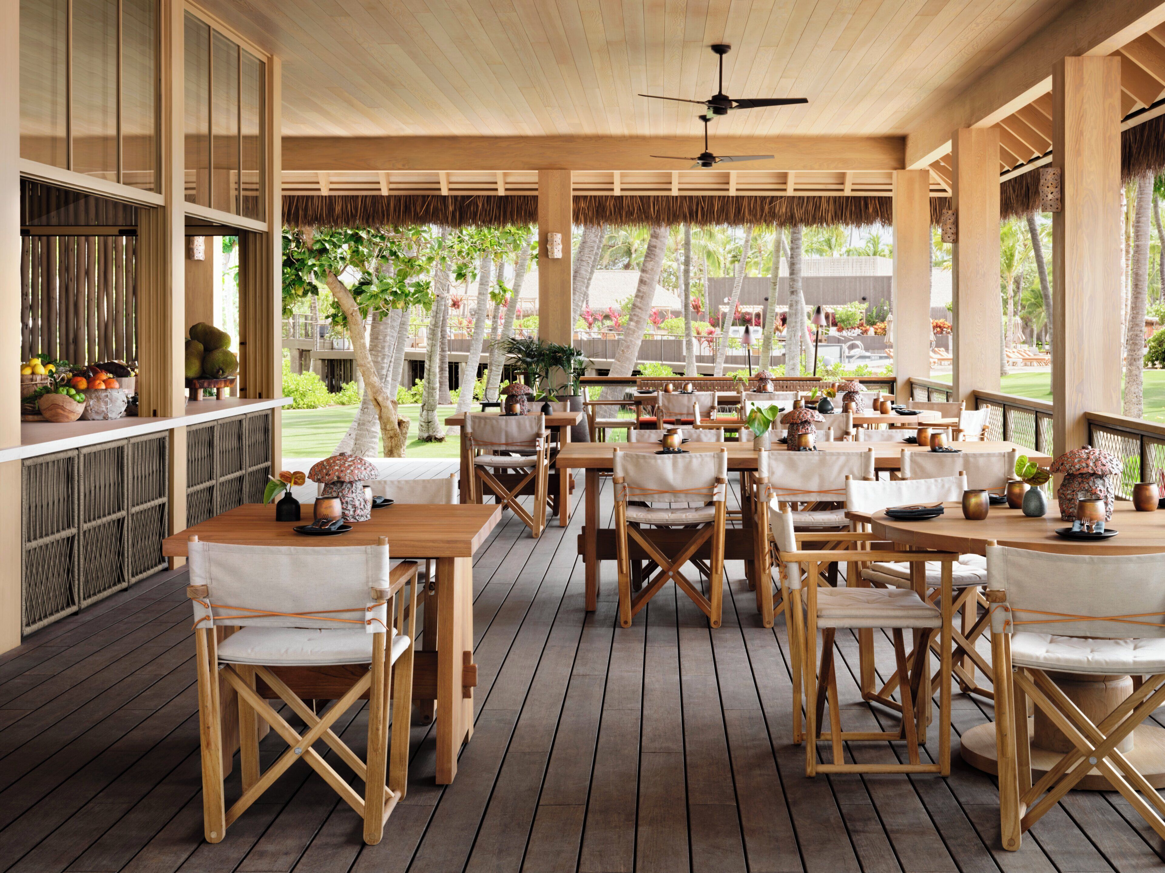 An outdoor dining area with wooden tables and chairs, a ceiling fan, and a view of a landscaped garden.