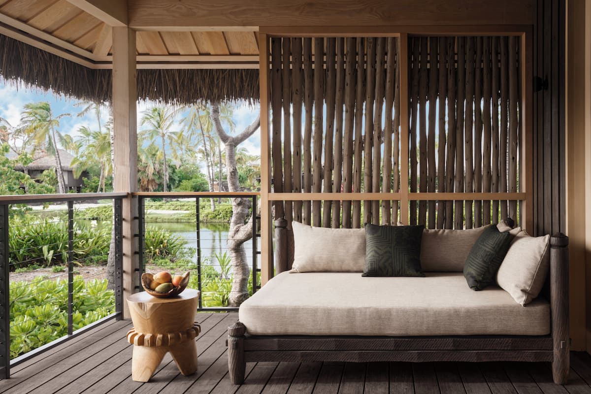 A wooden deck with a sofa and a small table, overlooking a pond and palm trees.