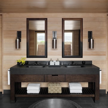 A modern bathroom with a wooden wall, two mirrors, a black sink, and a towel rack.