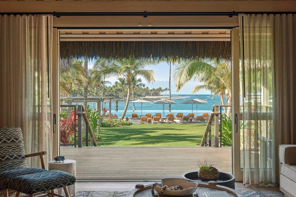 A room with a view of a pool and beach, featuring a wooden floor, a chair, a small table, and a bowl.