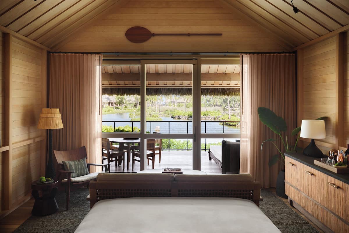 A room with a large bed, a dining table with chairs, and a view of a water body and greenery outside.
