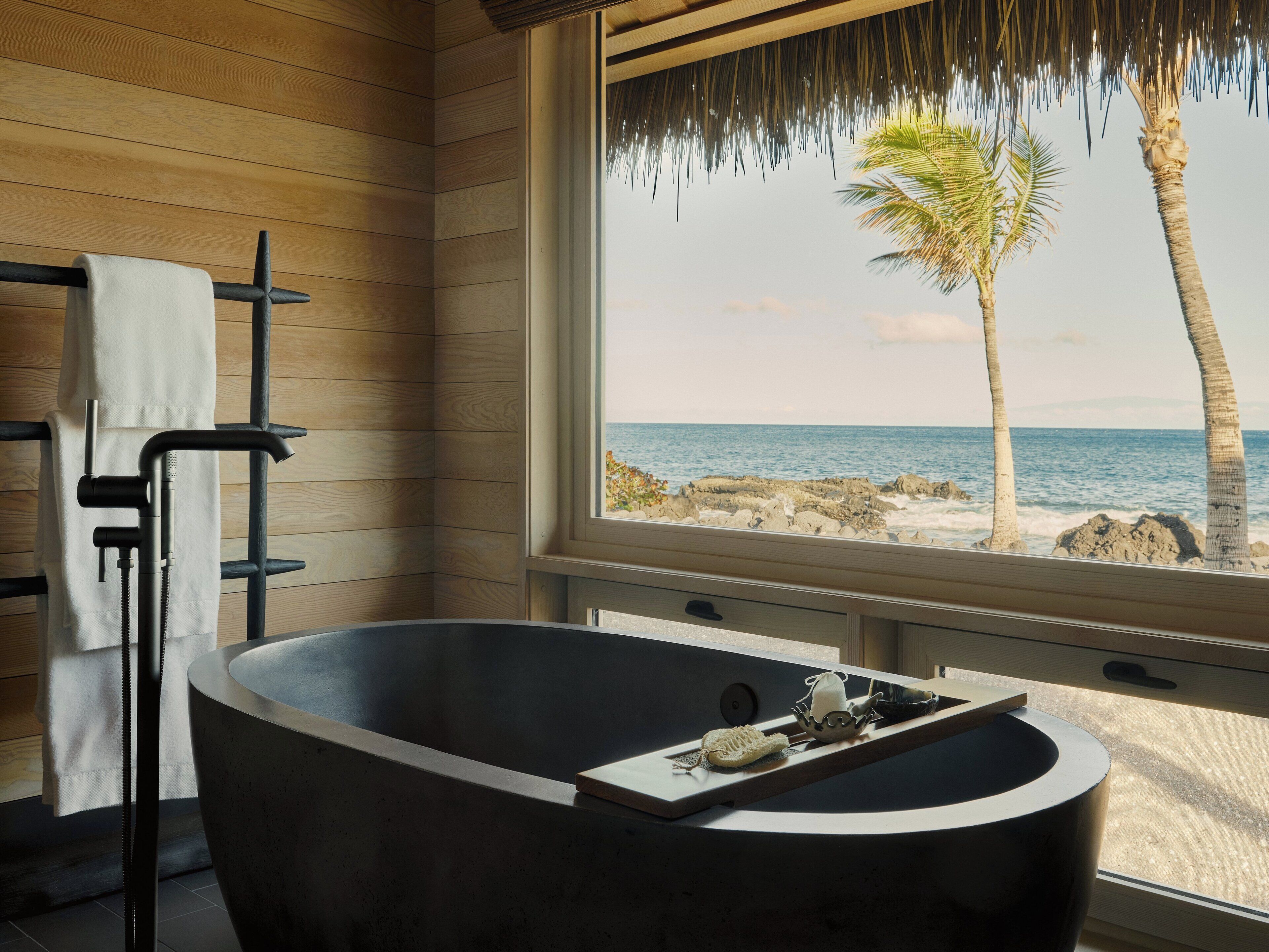 A modern bathroom with a freestanding bathtub, wooden panel walls, and a view of the ocean through a large window.