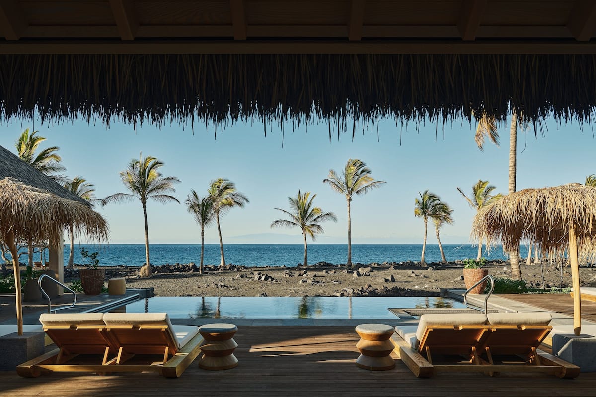 A wooden deck with lounge chairs and a pool, overlooking a beach with palm trees and a clear blue sky.
