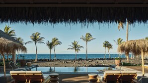 Une terrasse en bois avec des chaises longues et une piscine, des palmiers et une plage en arrière-plan.