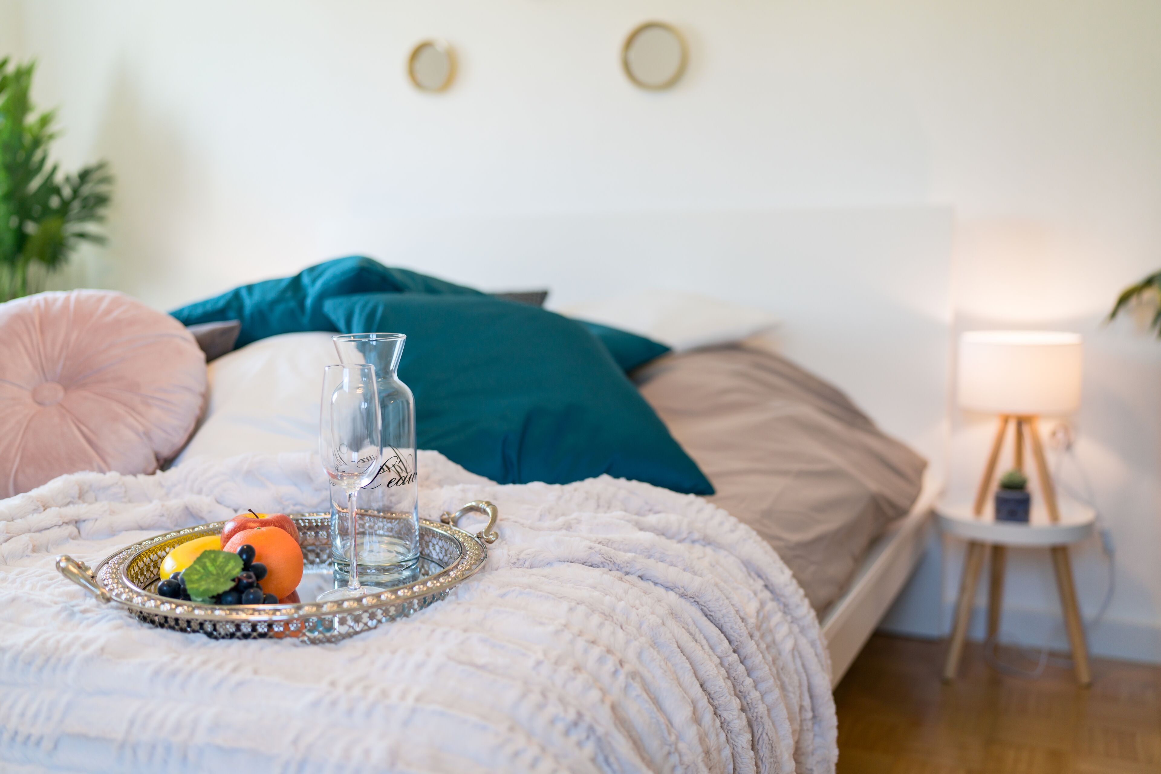 Una cama bien hecha con una bandeja decorativa que contiene frutas y un vaso de agua, una lámpara de noche y una planta pequeña en una mesita.