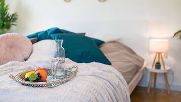Una cama bien hecha con una bandeja decorativa que contiene frutas y un vaso de agua, una lámpara de noche y una planta pequeña en una mesita.