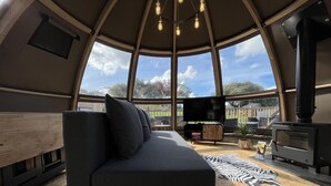 A cozy interior with a sofa, a television, a fireplace, and a large window offering a view of the outdoors. - Sunrise Dome Tent (Tenby)