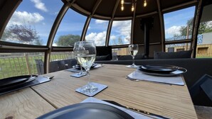 A dining table set for two with wine glasses, plates, and cutlery, in a room with a curved ceiling and large windows. - Sunrise Dome Tent (Tenby)