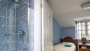 A bedroom with a shower, a bed with a checkered bedspread, a nightstand with a lamp, and a window with curtains. - Penybont Apartment (Aberystwyth)
