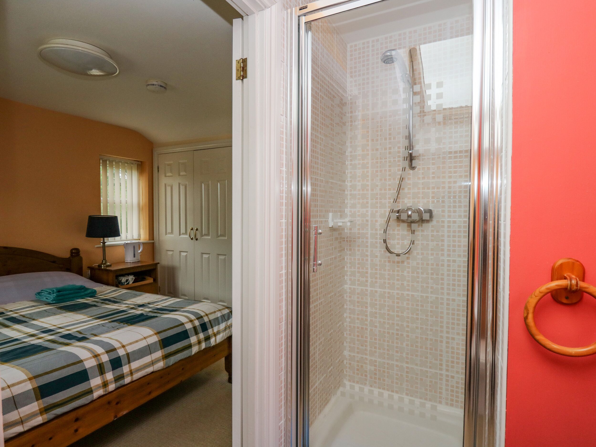 Un dormitorio con cama, mesita de noche, lámpara, ventana con persianas, puerta y zona de ducha con puerta de cristal.