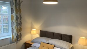 A bedroom with a bed, two bedside tables, a window with curtains, a hanging light fixture, and a radiator. - Hursley Lodge (Lincoln)