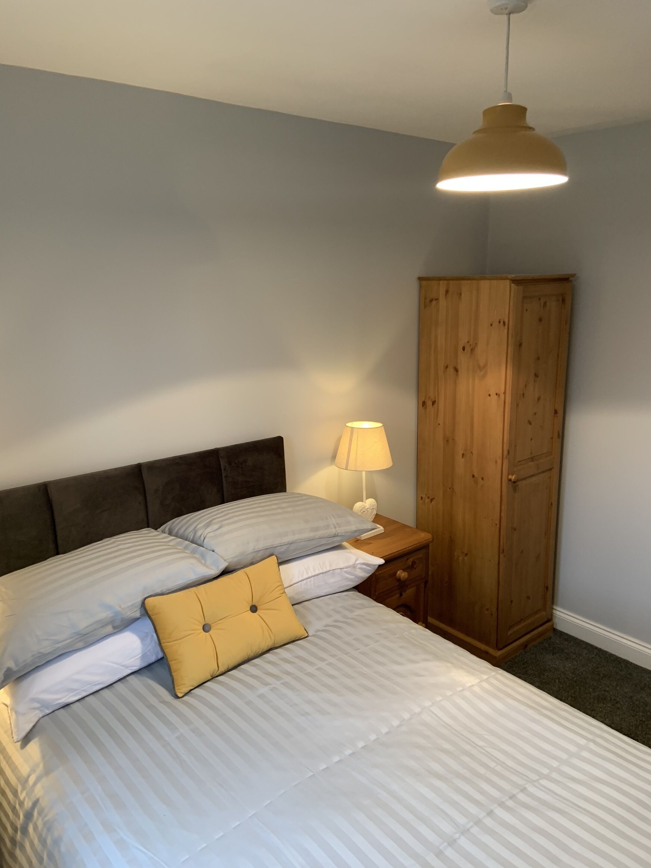 A bedroom with a bed, pillows, a nightstand with a lamp, and a wooden wardrobe.