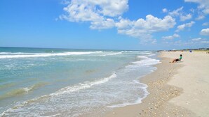 Ein Strand mit einer Person, die auf einem Stuhl sitzt, sanften Wellen, die ans Ufer schlagen, und einem klaren Himmel mit vereinzelten Wolken.