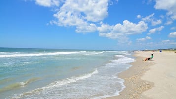A beach with a person sitting on a chair, waves gently lapping the shore, and a clear sky with scattered clouds.