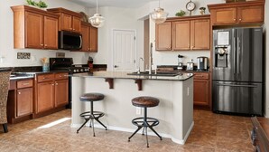 A modern kitchen with wooden cabinets, a stainless steel refrigerator, a central island with stools, and pendant lighting. - Estrella Golden Oasis | Mountain view back yard! (Goodyear)