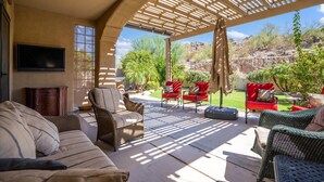 A covered patio area with wicker furniture, a flat-screen TV, and a view of a landscaped garden. - Estrella Golden Oasis | Mountain view back yard! (Goodyear)