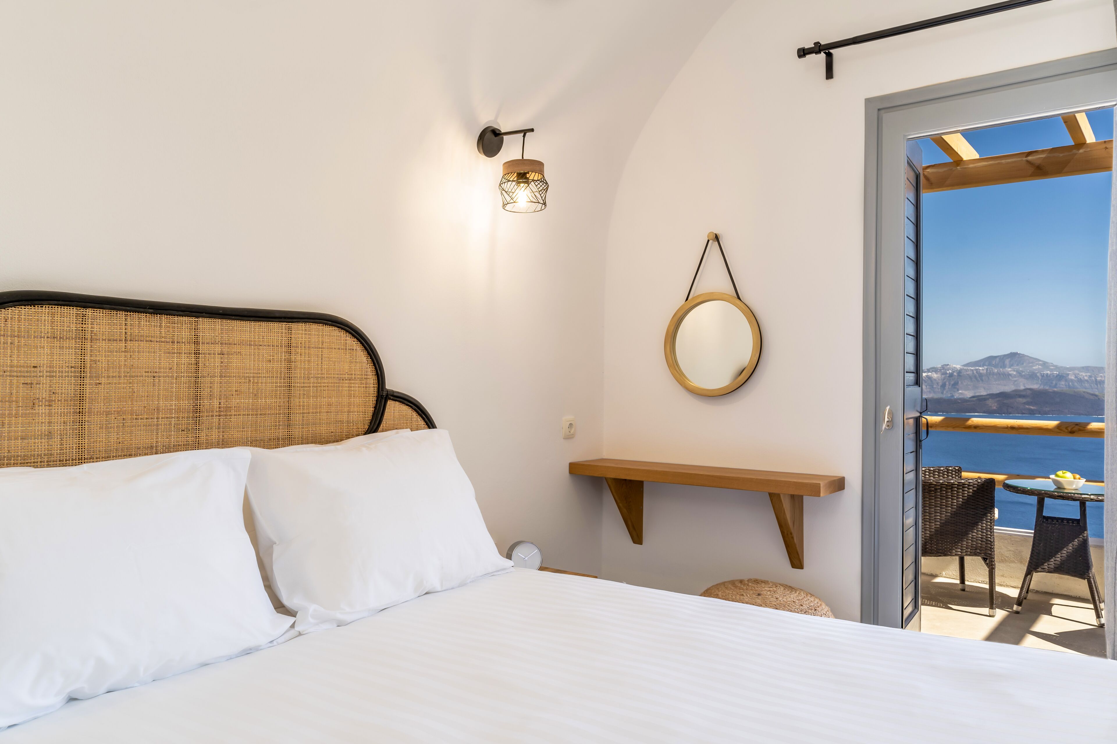 A bedroom with a bed, a wall-mounted light, a wooden shelf, a round mirror, and a balcony with a view of the sea.