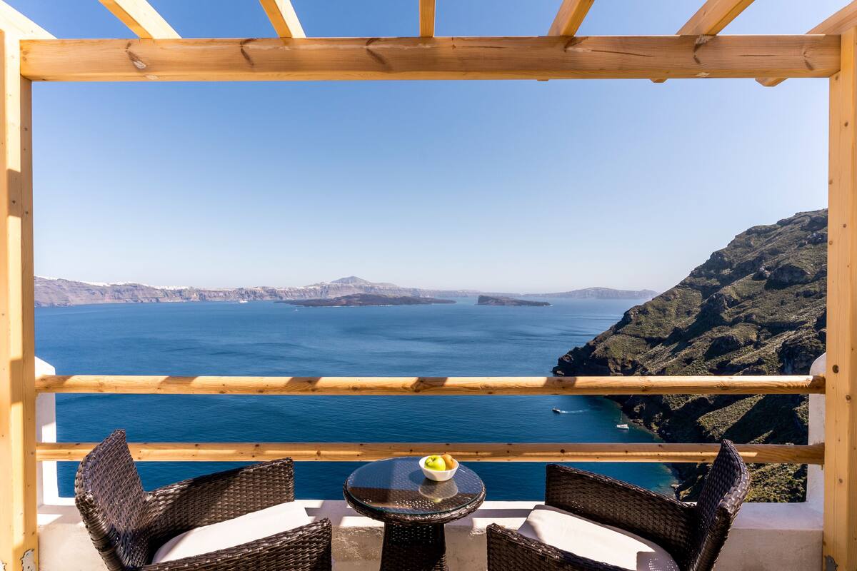 A balcony with wicker furniture and a table with a bowl of fruit, overlooking a coastal landscape with clear blue sea and distant mountains.